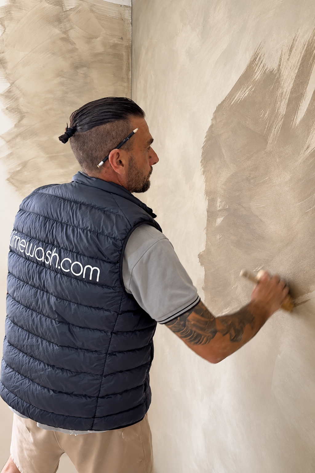 A man wearing a navy blue puffer vest with 'crewall.com' printed on the back, applying a beige plaster or stucco wash to a wall in an indoor setting.