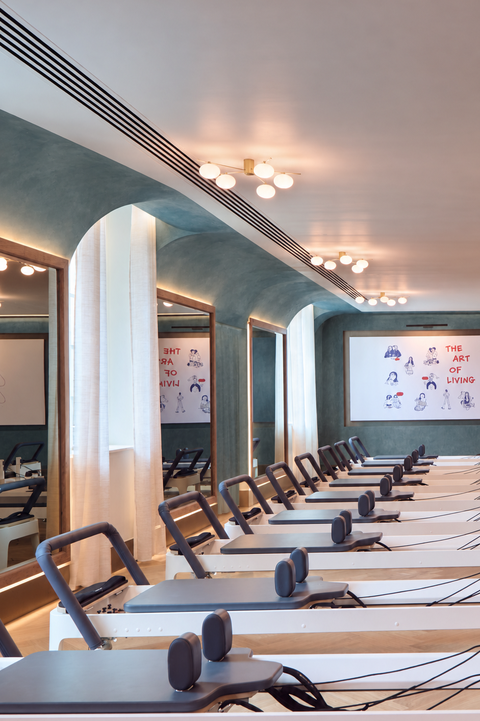 A row of Pilates reformer machines arranged in parallel rows inside a modern fitness studio with teal walls, arched doorways, and warm ceiling lights.