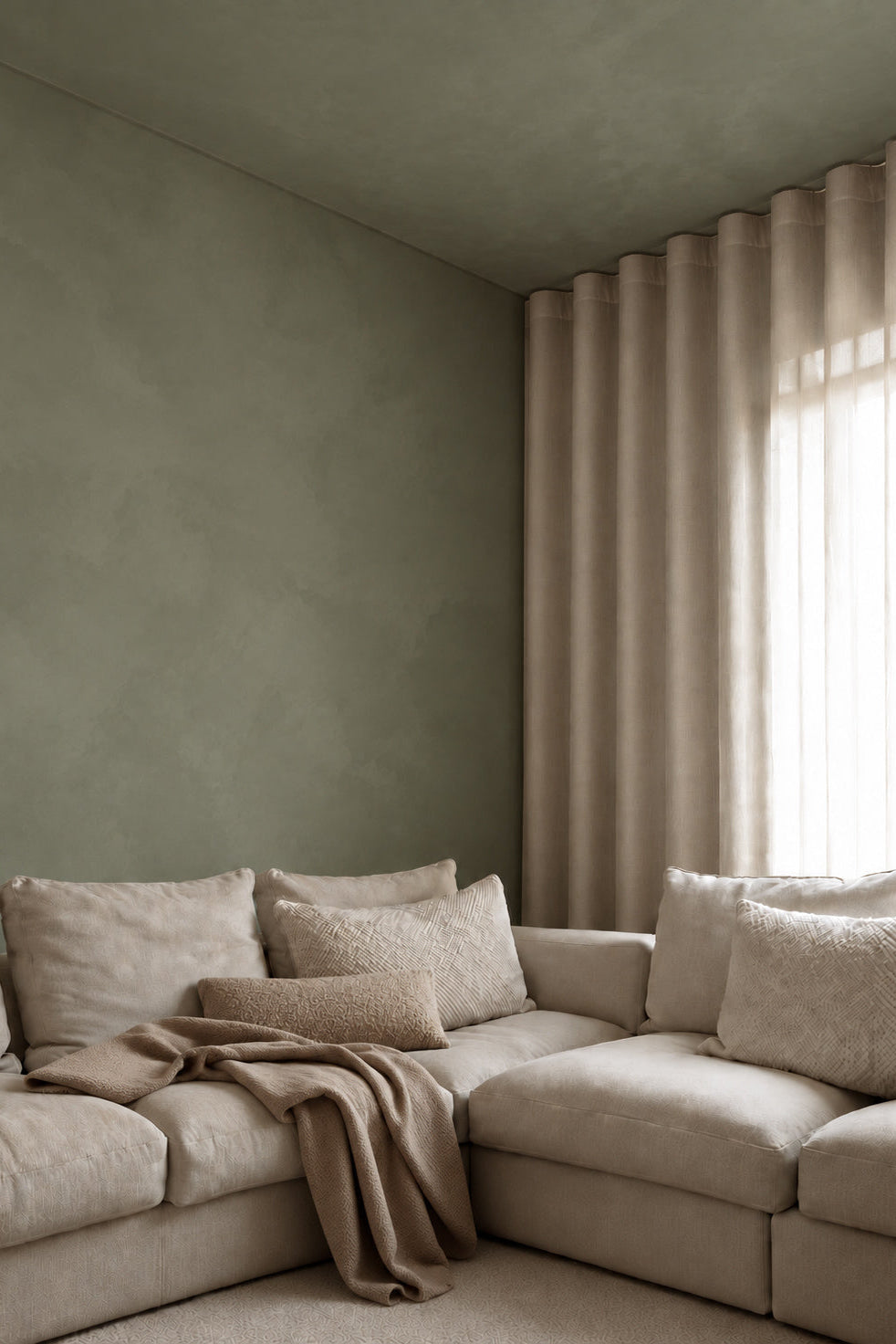 A styled living room corner featuring a beige sectional sofa with textured throw pillows and a draped throw blanket, set against a Limewash sage green wall and a window with beige pleated curtains.