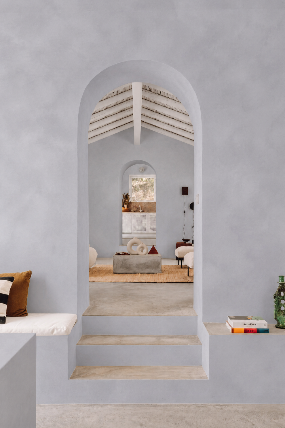 A modern interior design scene featuring a light gray stucco wall with a large arched doorway looking into a bright room with white walls, a concrete coffee table, and a vaulted ceiling. In the foreground, a matching light gray bench with a striped pillow is visible on the left, and a green decorative bottle and books sit on a shelf on the right.