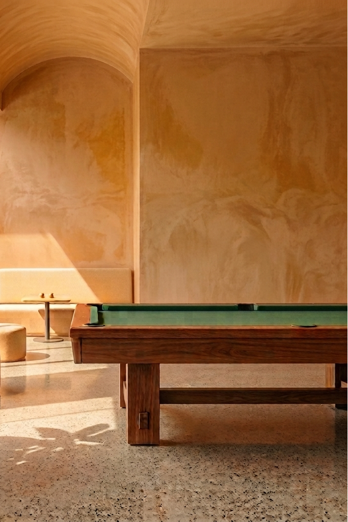 A modern interior scene with a wooden pool table featuring a green felt surface, set in a warm, sunlit room with terracotta walls, terrazzo flooring, and a large potted plant in the foreground.