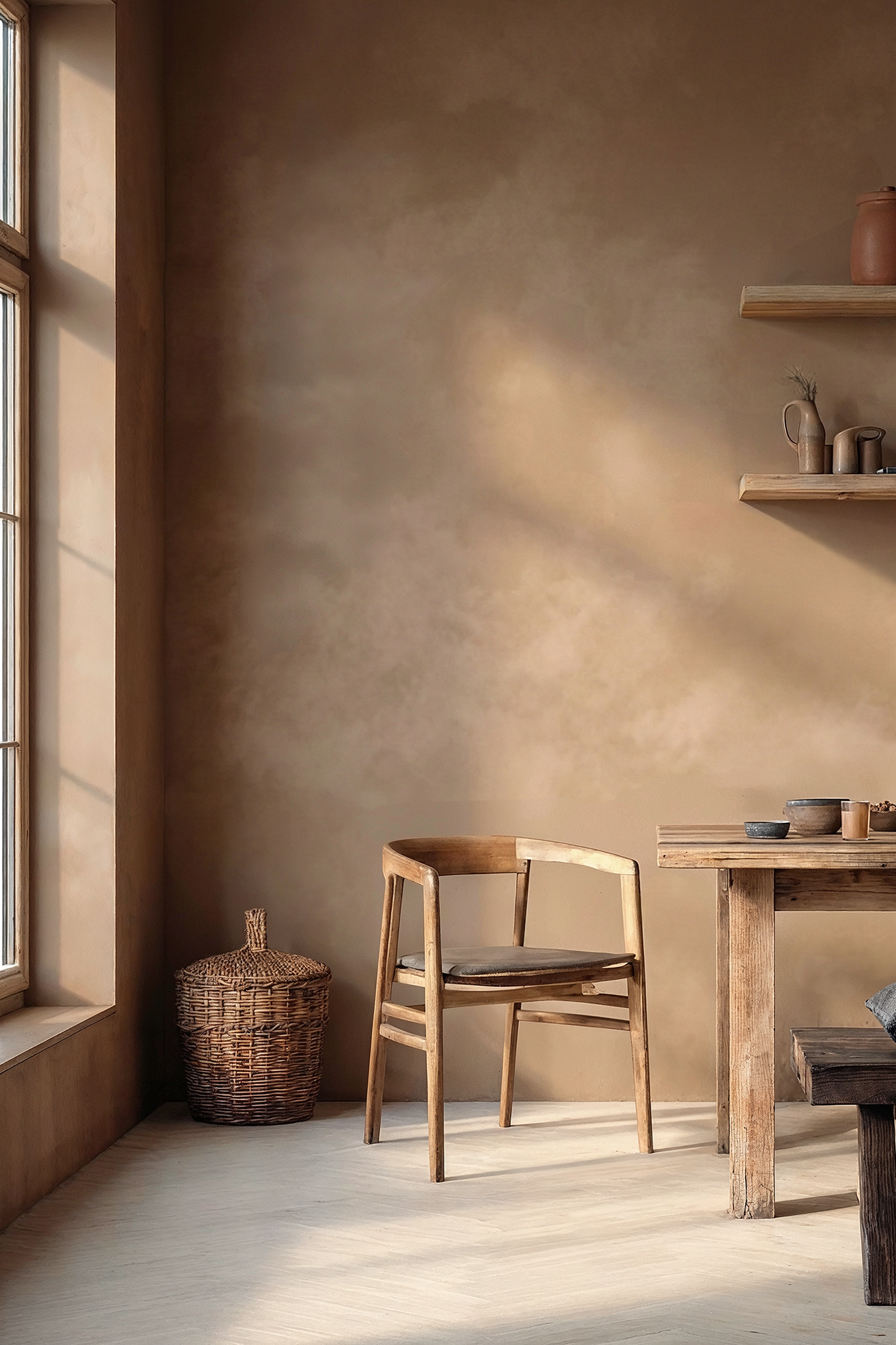 A warm, rustic interior scene with a wooden dining table, matching chair, bench with dark cushions, floating shelves, and a limewash walls with limewash.com brown  wall lit by sunlight from a window.