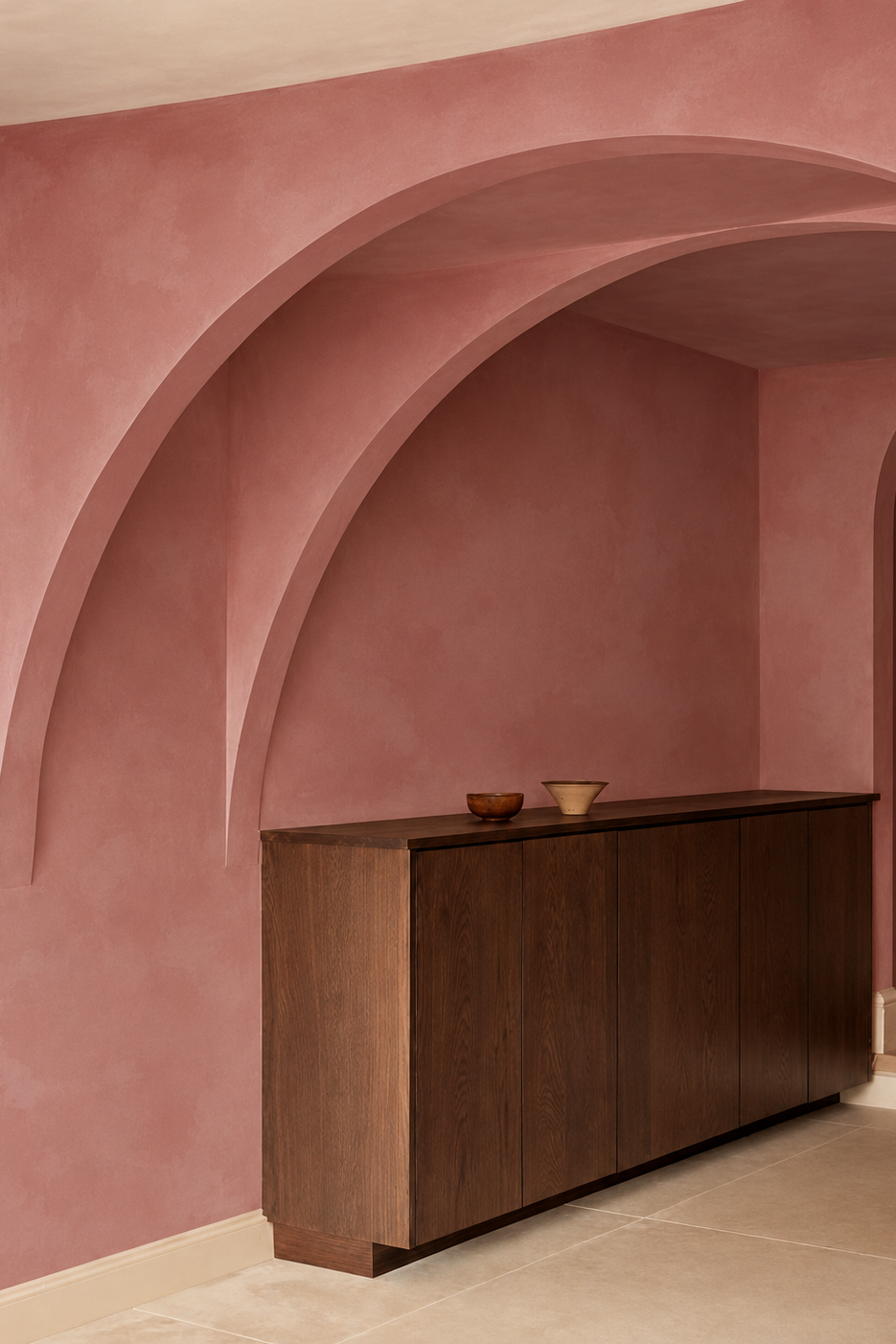 A modern dark wood sideboard cabinet with a minimalist design, set against a dusty rose plaster wall with arched architectural details.