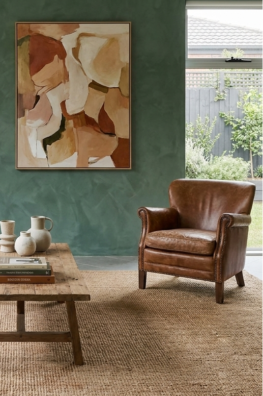 A styled living room interior with a green textured wall, a cream slipcovered sofa, a brown leather armchair, a wooden coffee table, a potted plant, an arched floor lamp, and a framed abstract painting.