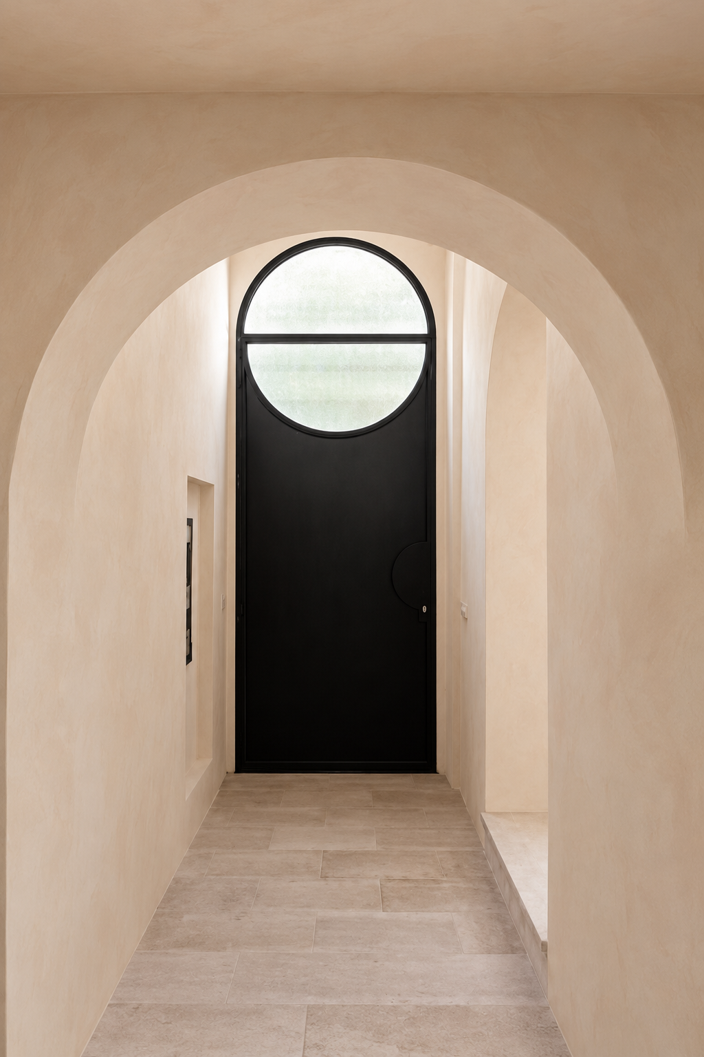 A modern black entryway door with a large circular window, set within a warm beige stucco hallway framed by an archway limewashed