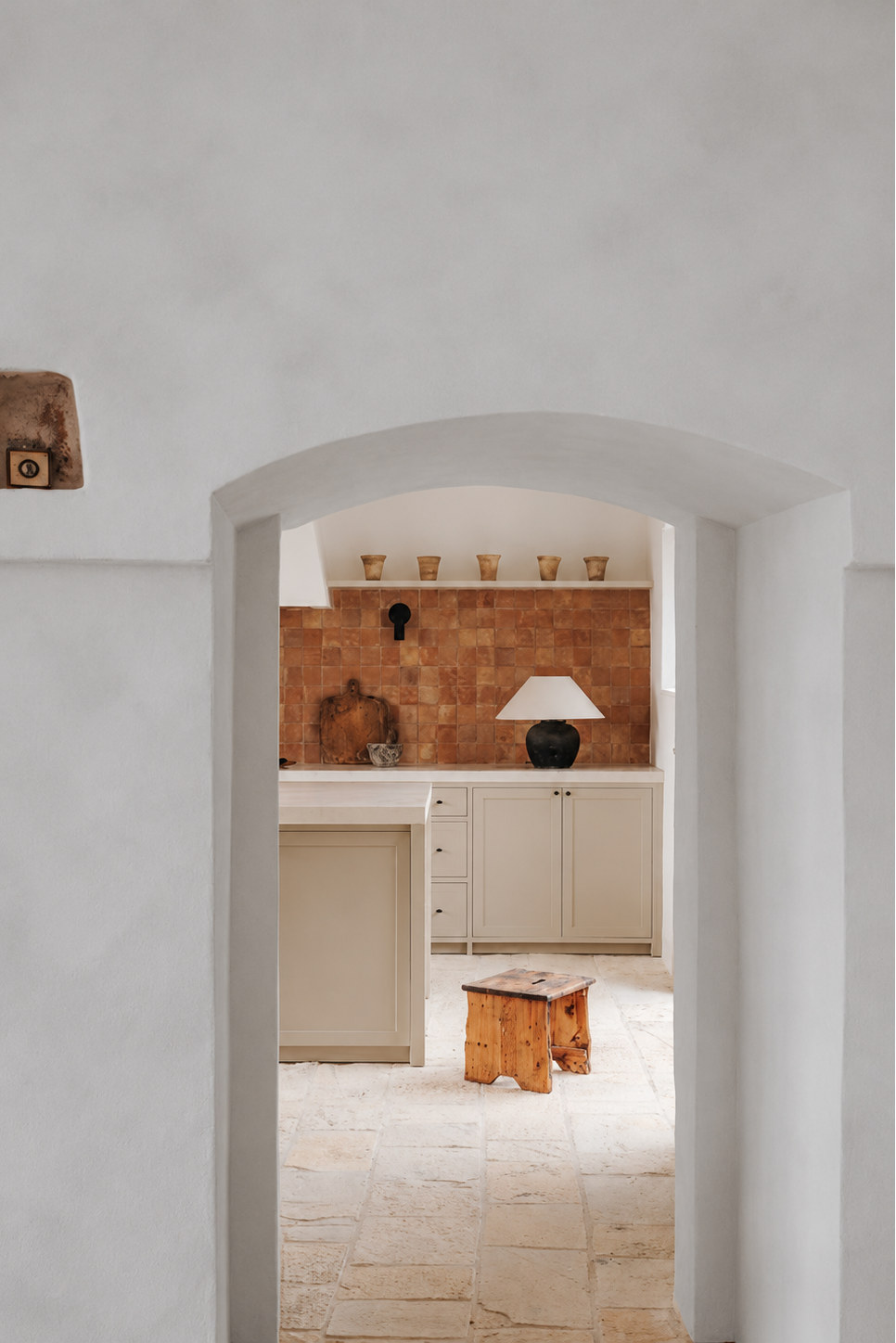 An interior architectural scene viewed through a white plaster archway, showing a rustic kitchen with terracotta tile backsplash, cream cabinetry, a small wooden stool, and stone flooring.