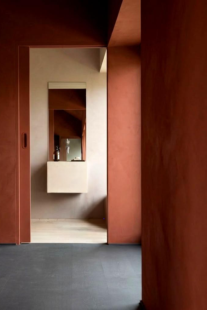A minimalist interior architectural scene with muted red -colored limewash walls, a dark gray floor, and a recessed doorway leading to a cream-colored vanity area with a mirror.
